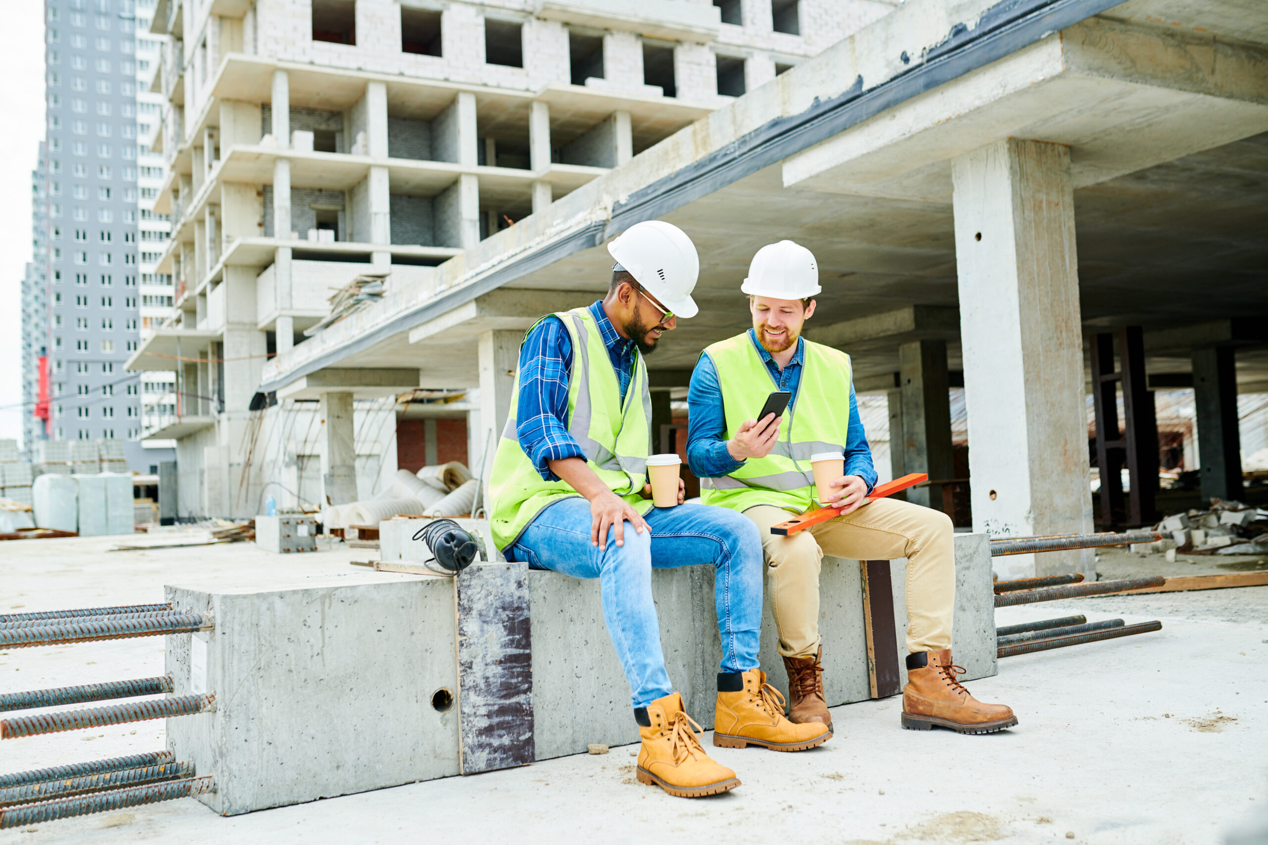 Full length portrait of two workers wearing hardhats chatting and using smartphone during coffee break on construction site