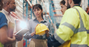 Logistics, team and safety meeting with helmet for PPE compliance, injury prevention and flare. People, warehouse supervisor and discussion with hardhat for OSHA regulations, hazard reporting or risk.